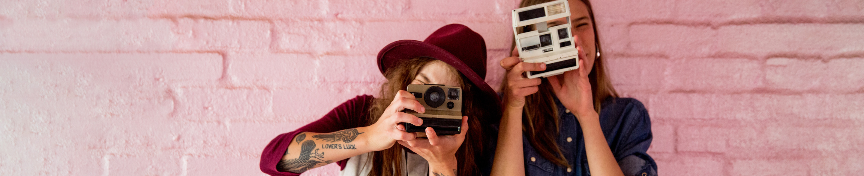 Dvě osoby drží retro fotoaparáty před růžovou cihlovou zdí.