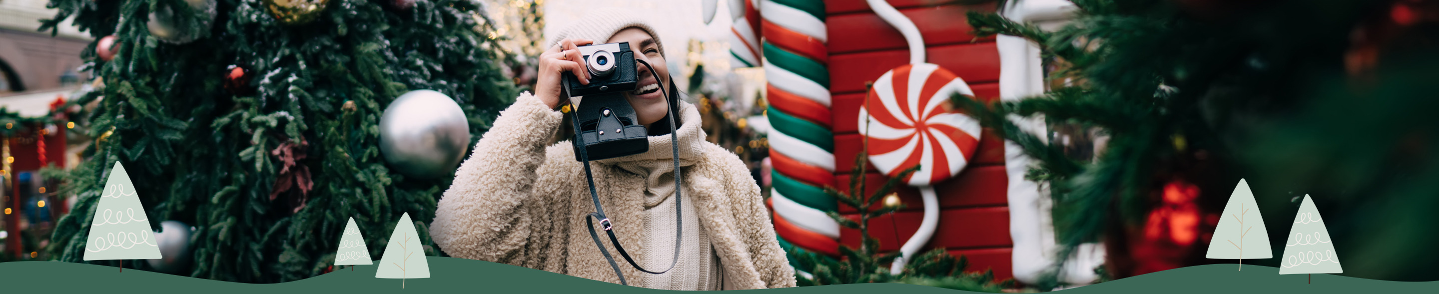 Osoba v béžovém kabátu drží fotoaparát před velkým ozdobeným vánočním stromem a červenobílými vánočními dekoracemi
